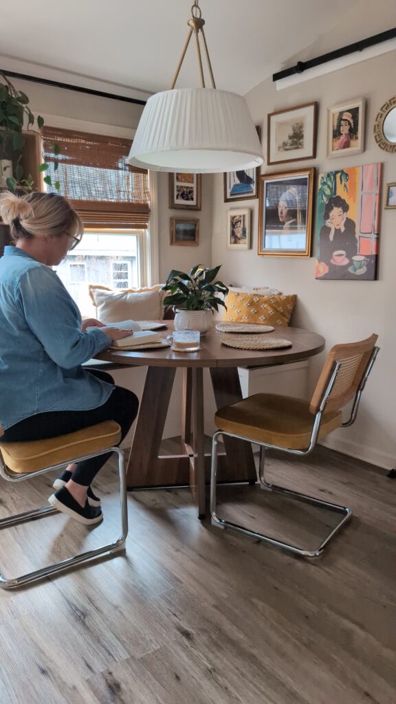 Woman seated at light and cozy breakfast nook featuring a round table, cane chairs, soft textiles, and a warm gallery wall.