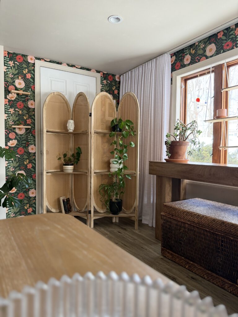 Collected living room corner with rattan folding screen, indoor plants, floral wallpaper, and warm wood furniture
