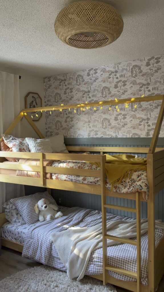 Cozy kids shared bedroom with wooden bunk bed, warm string lights, floral wallpaper and layered neutral bedding.