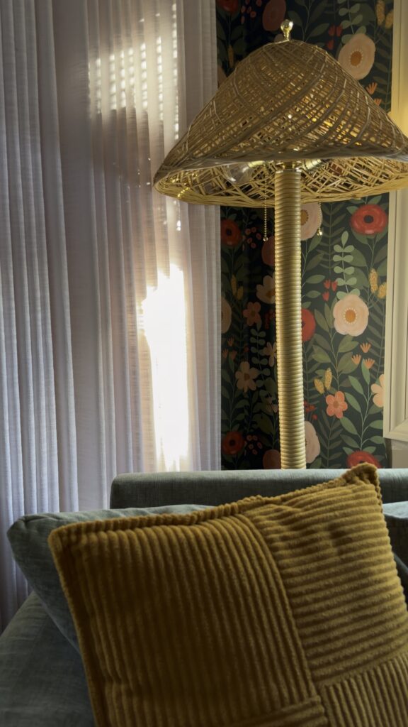 Woven rattan floor lamp glowing beside green sofa and patterned wallpaper in warm neutral living room.