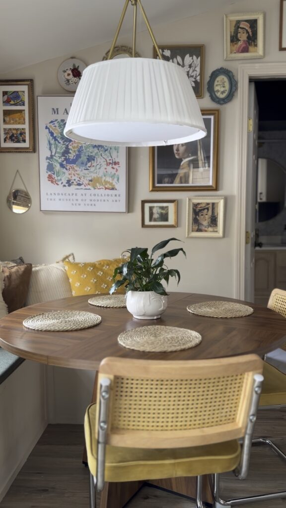 Pleated white pendant light over round wood dining table in cozy breakfast nook with gallery wall and cane chairs.
