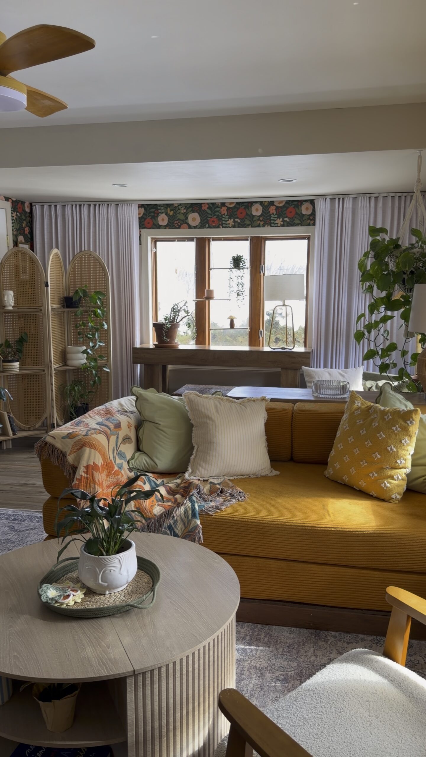 Cozy living room with mustard sofa, floral wallpaper, layered pillows, warm lighting, and round wood coffee table.