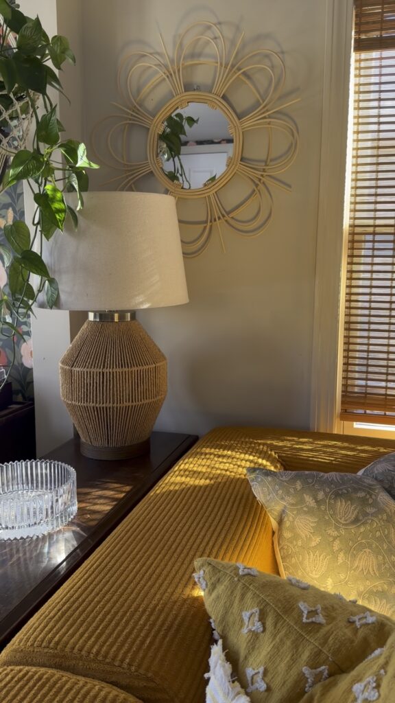 Boho rattan mirror above a textured table lamp and mustard sofa with patterned pillows in a cozy living room