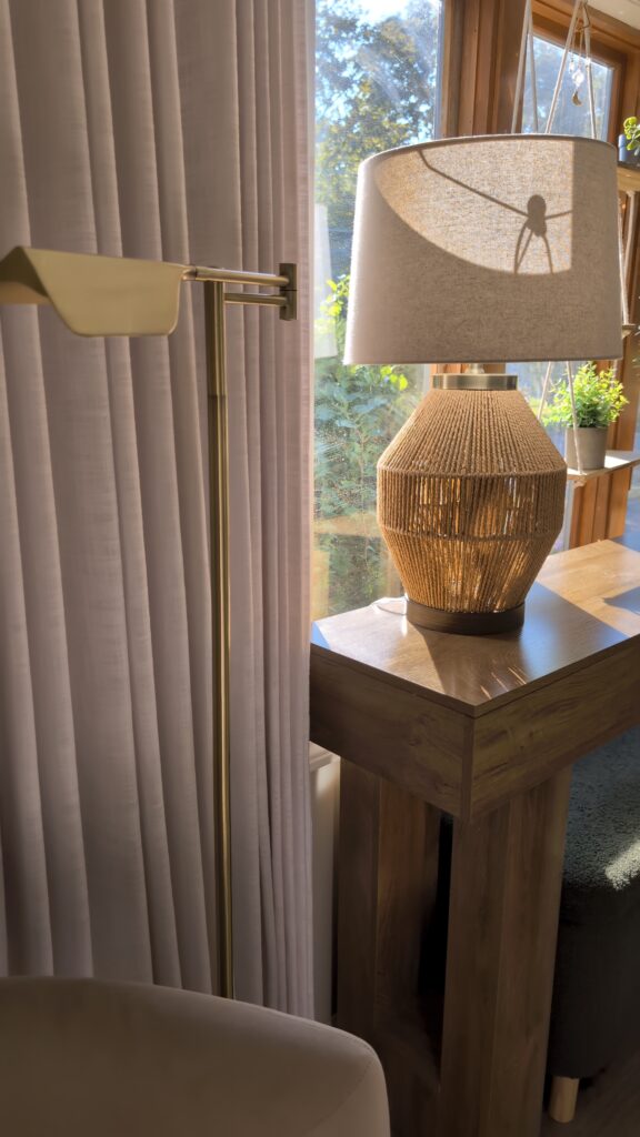Cozy living room corner with brass floor lamp and woven textured table lamp on wood side table near sheer curtains.