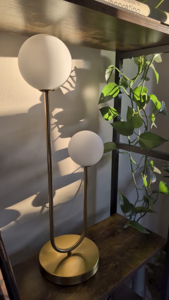Double globe modern table lamp with warm lighting styled on dark wood shelving next to trailing houseplant.
