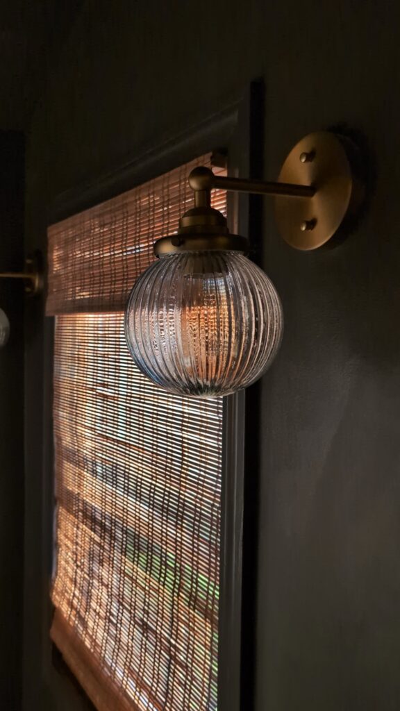Warm brass bathroom wall sconce with ribbed glass globe casting soft light against dark green walls and woven window shade.
