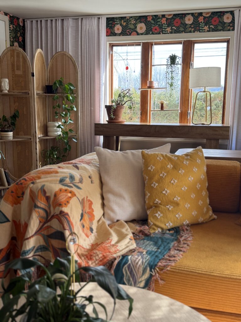 Mustard sofa layered with neutral and patterned pillows in front of floral wallpaper and soft linen curtains.