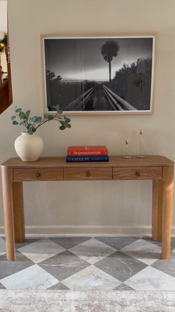 light wood console table styled under framed wall art in entryway
