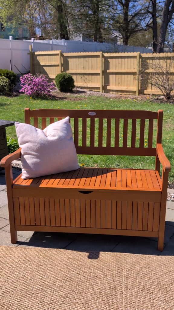 Wood outdoor storage bench on a backyard patio styled with neutral outdoor pillows.
