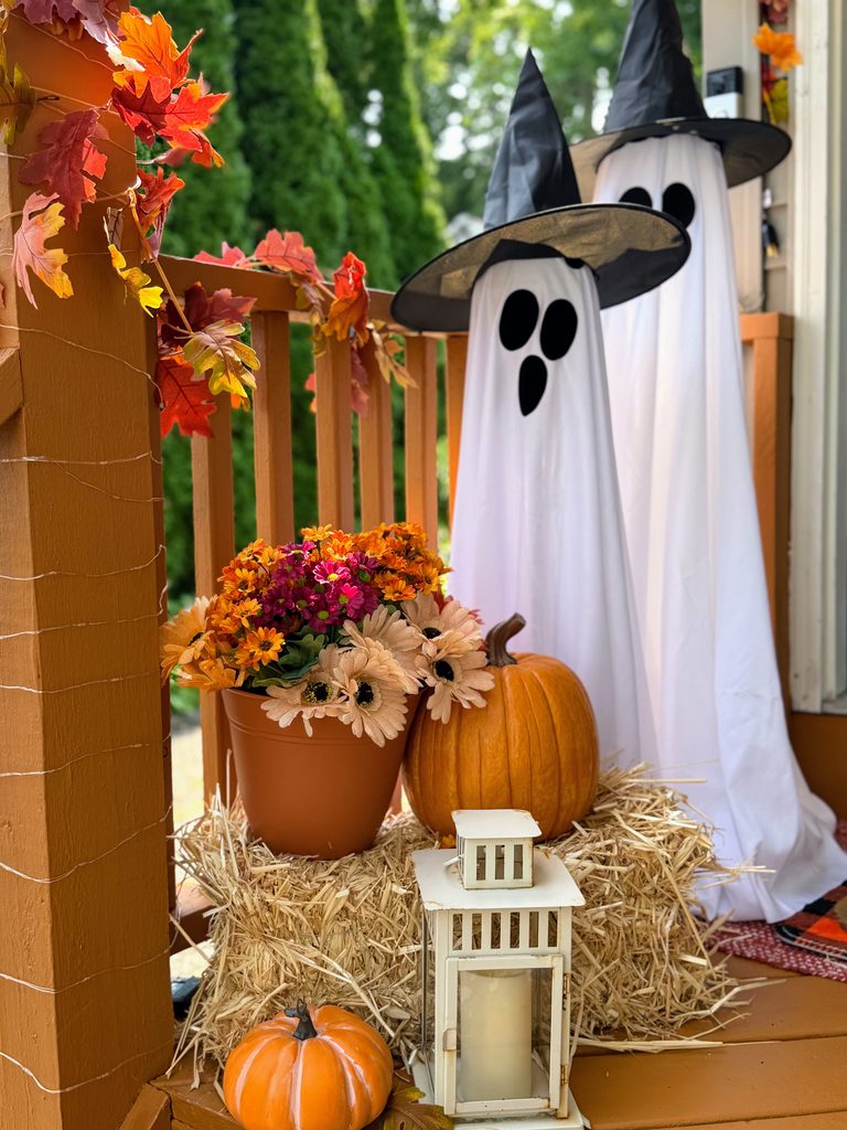 White Halloween witch ghosts displayed with pumpkins, fall flowers and a bale of straw on a front porch
