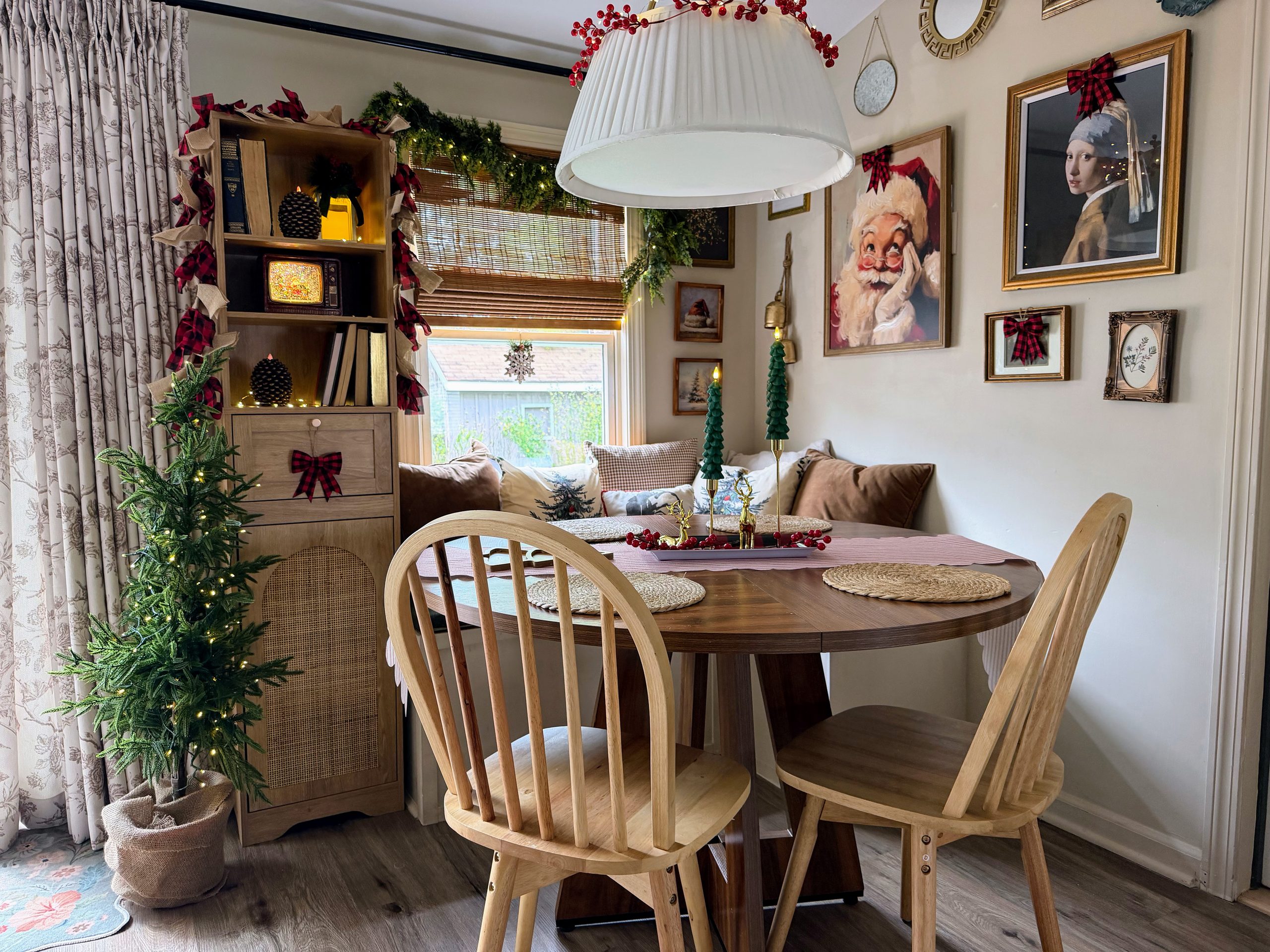 Cozy Christmas breakfast nook with vintage Santa artwork, festive garland, plaid bows, a small lighted Christmas tree, and a round wooden dining table decorated with Christmas tree candles.