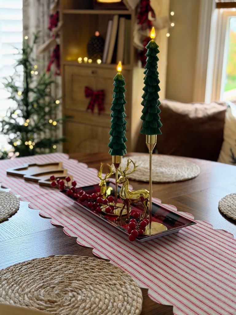 Green Christmas tree taper candles with brass holders, gold reindeer, and red berries on a striped scalloped table runner.