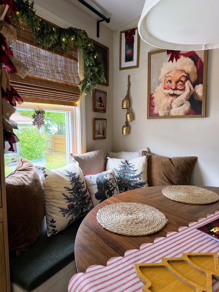 Cozy window seat with Christmas tree and Santa pillows, brown cushions, bamboo shades, and lighted evergreen garland.