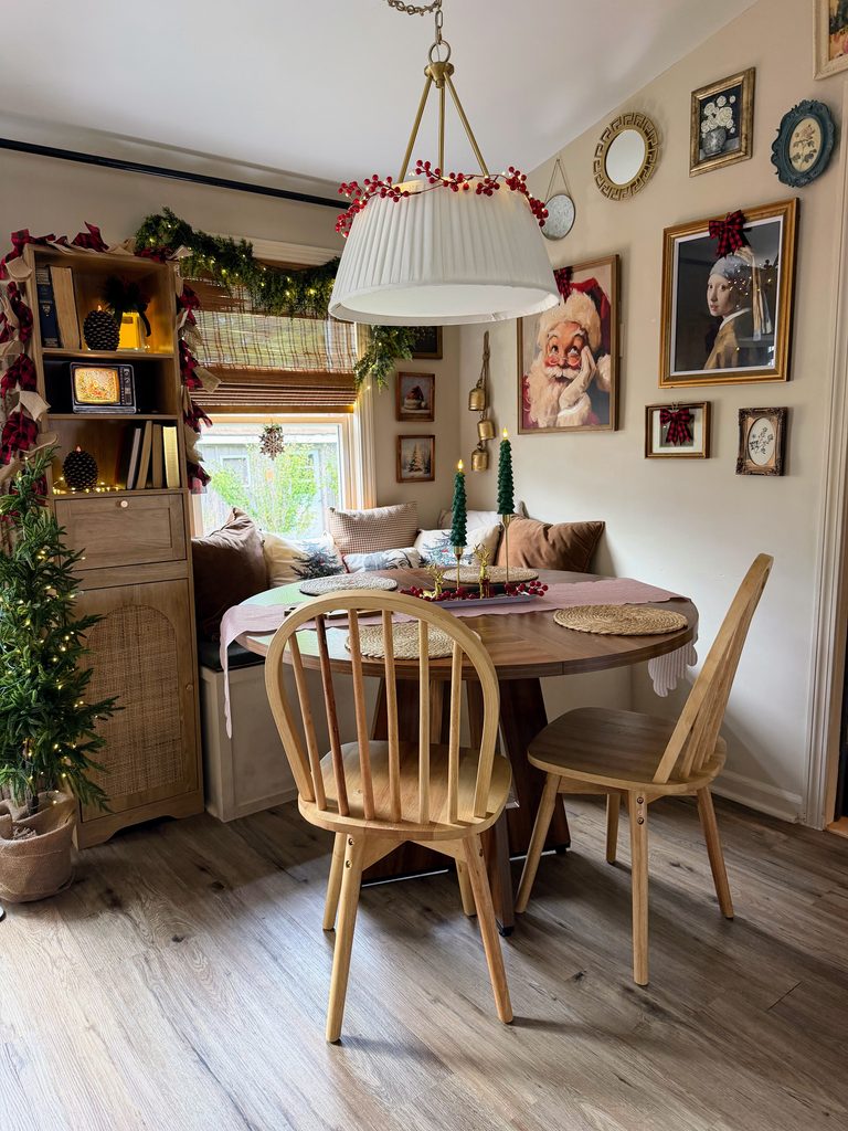 Small Christmas breakfast nook decorated with a window bench, holiday pillows, greenery, festive artwork, and a wooden dining table.