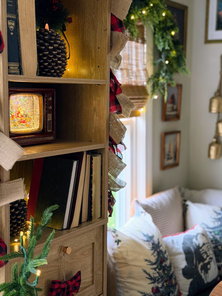 Vintage television-shaped Christmas snow globe glowing on a wooden bookshelf with pinecone candles, books, and plaid ribbon garland.